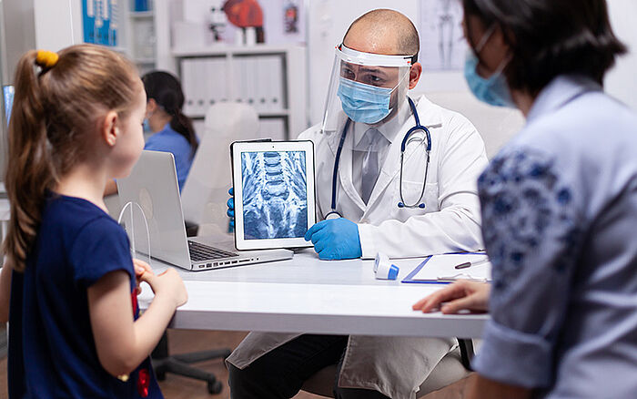 A close-up of a young girl speaking with a pediatrician, both wearing masks, during a medical consultation in a hospital office.