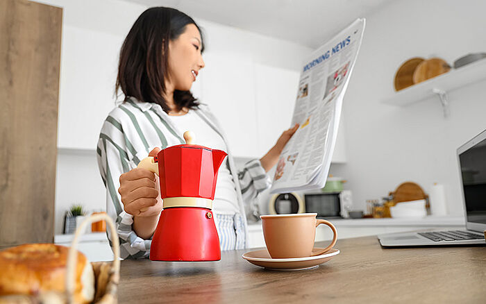 A woman in pajamas lifts a red moka pot to pour herself a coffee while reading a newspaper.