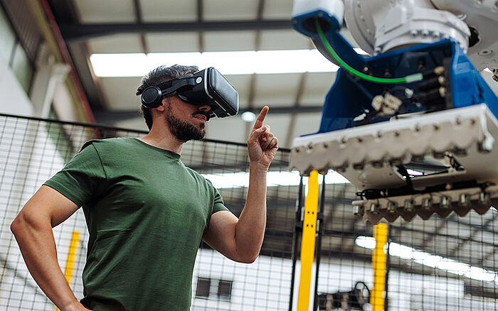 Industrial engineer wearing a virtual reality headset while controlling a robotic arm in a high-tech factory.