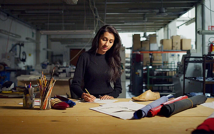 Designer sketching footwear designs at a worktable in a studio, with fabric rolls and sample shoes nearby.