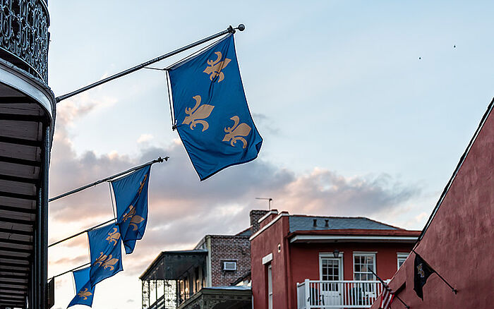 View of Bourbon Street in New Orleans' historic Old Town at sunset, featuring classic balcony architecture and blue flags hanging from wrought-iron railings under a dramatic, cloud-filled sky.