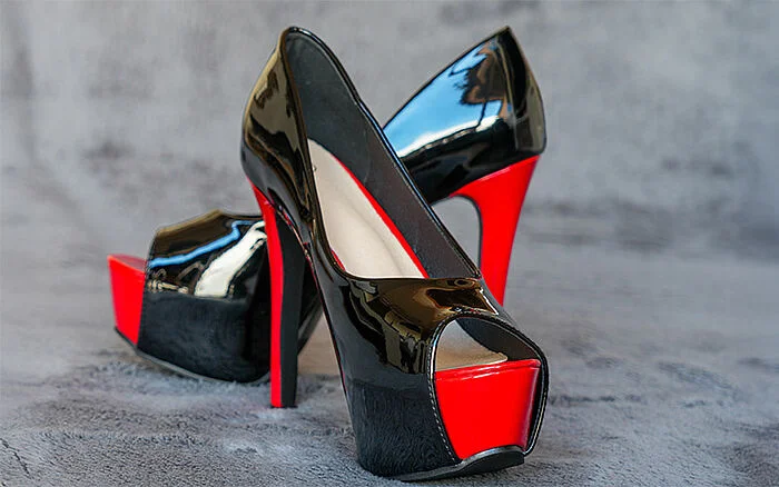 Glossy black high-heeled platform shoes with bright red soles and accents, positioned on a gray surface.