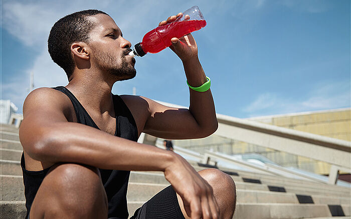 Handsome African runner with muscular arms and legs sitting on concrete steps after a run, refreshing and quenching his thirst by drinking a juice or shake from a plastic bottle.