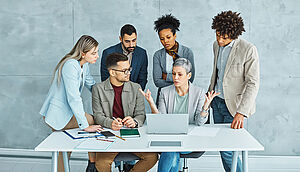 Young business professionals in a modern office engaged in a team meeting. A woman presents information using a laptop, while colleagues seated at a desk listen attentively, suggesting a collaborative startup or training environment.