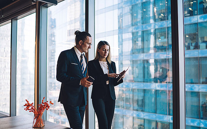 Mature man and woman in business attire examining financial data on a digital touchscreen tablet and desktop display, conveying professional data review and information analysis.