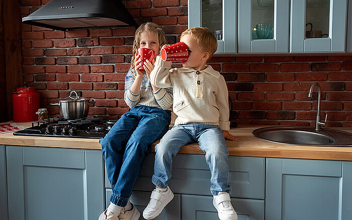A young brother and sister have fun sitting on a kitchen countertop, drinking from oversized mugs.