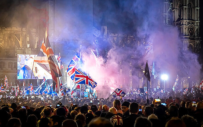 People celebrating Brexit in Parliament Square, London, with Union Jack flags and political signs in the background.