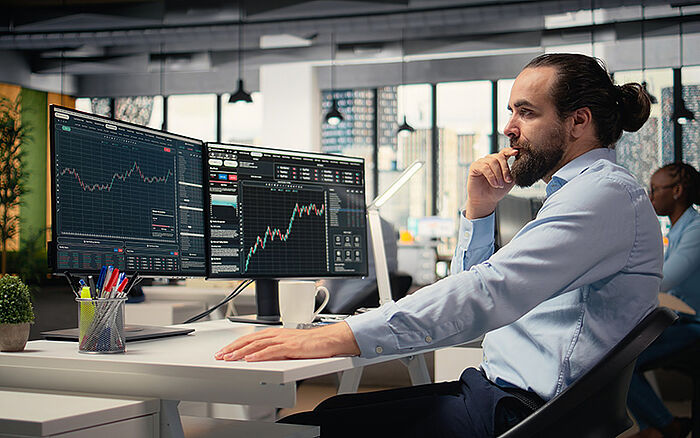A financial professional in an office setting looks at multiple computer screens showing stock exchange data.