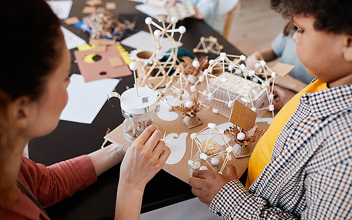 High-angle close-up photo of a young boy showing a school project (art & craft) to a female teacher, inside a classroom.