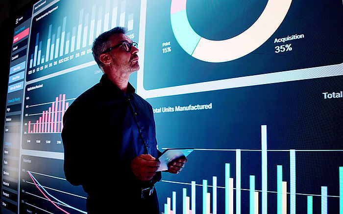 Man holding a tablet looks at a large wall display of business analytics charts and graphs in a dark, high-tech office.