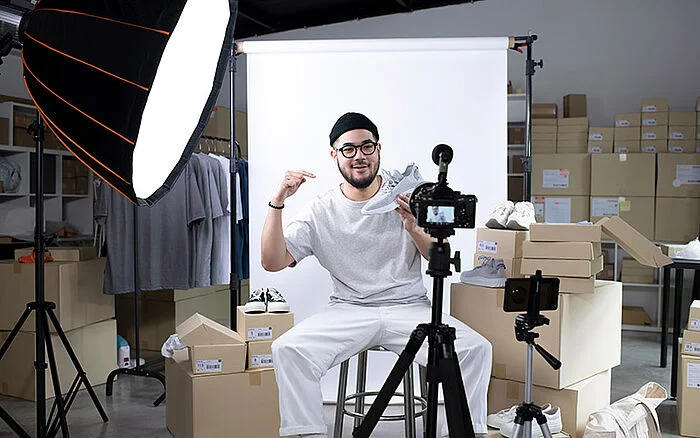 A young Asian man sits in a well-lit indoor setting, engaging with the camera while presenting a product, suggesting he is recording content for his social media or online business.