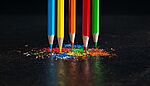 Five color pencils balance on their tips atop colored shavings on a black surface against a black background.