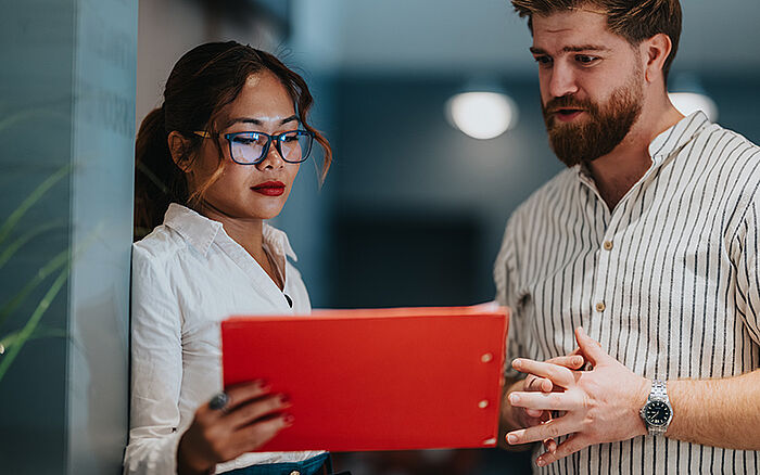Two professionals reviewing documents in a red folder at a desk in a modern office, engaged in focused discussion and collaboration.