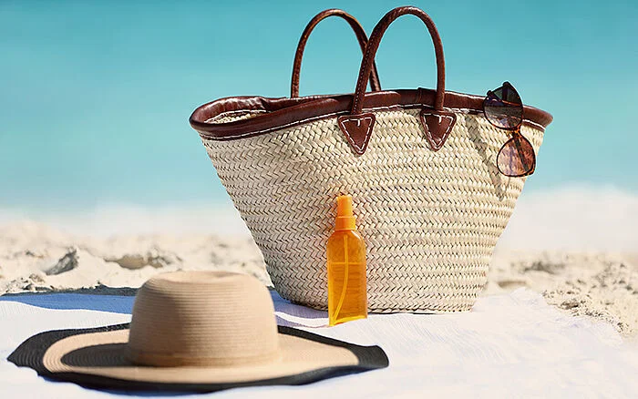 Women&rsquo;s beach accessories including a straw tote bag, sun hat, and sunscreen bottle on sand with a blue ocean in the background.