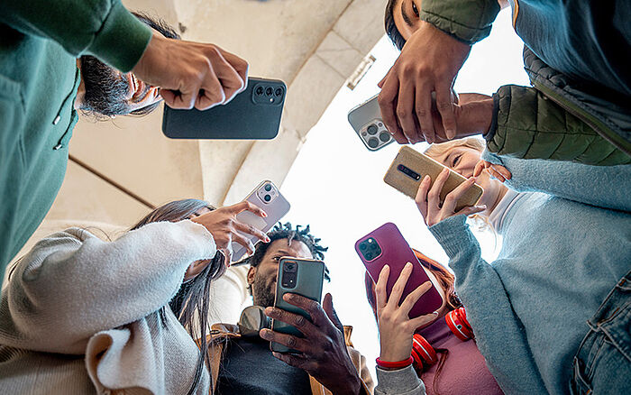 Group of young adults standing in a circle, viewed from below, each looking at a smartphone.