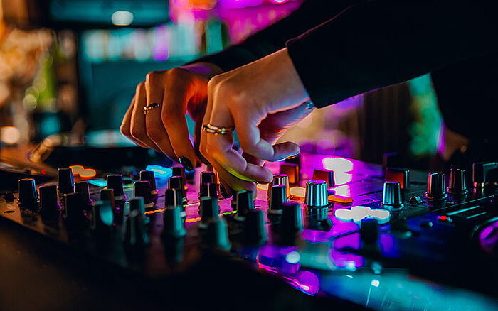 DJ adjusting music on a console mixer during a live performance in a nightclub, with hands on controls and colorful stage lighting.