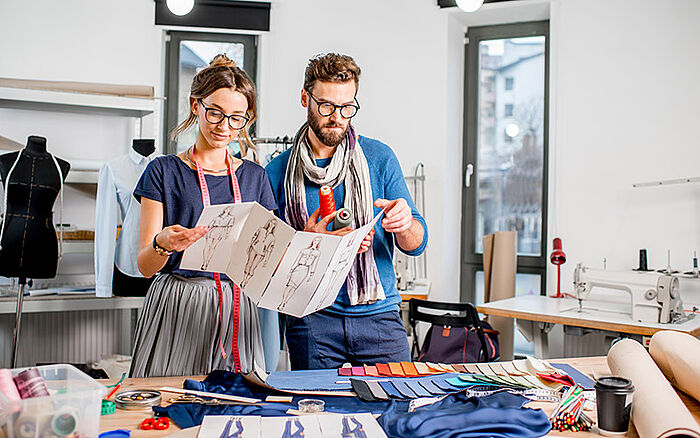 A male and female fashion designer working together in a bright studio as they review clothing sketches on a table filled with fabric samples and tailoring tools. They appear focused and creative, immersed in the design process.