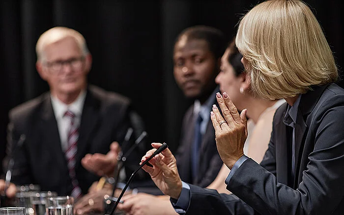 Rear view of a woman speaking into a microphone during a press conference or political debate while addressing a panel of officials in a conference room.