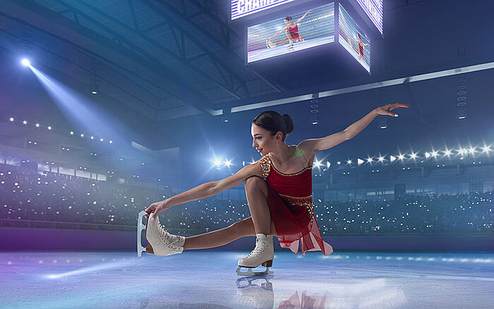 Young female figure skater gliding gracefully on the ice in a large indoor ice arena, wearing a skating outfit, with the smooth ice surface and arena lighting visible in the background.