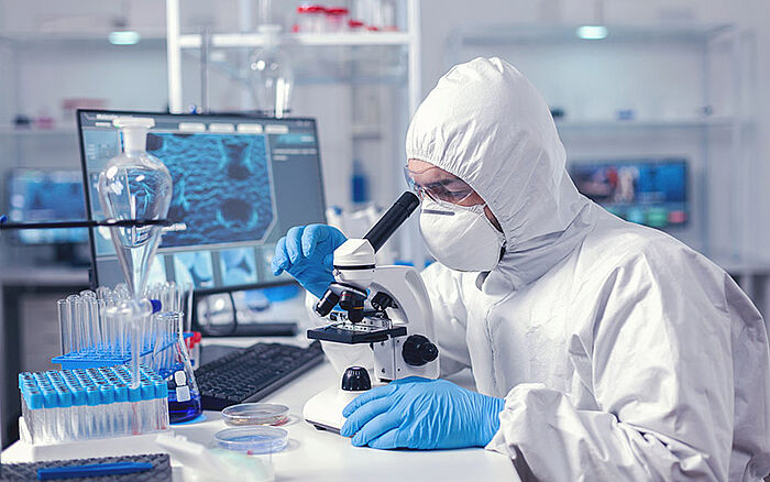 Biochemist in full PPE suit analyzing a sample under a microscope in a laboratory, conducting scientific research in a controlled healthcare environment.