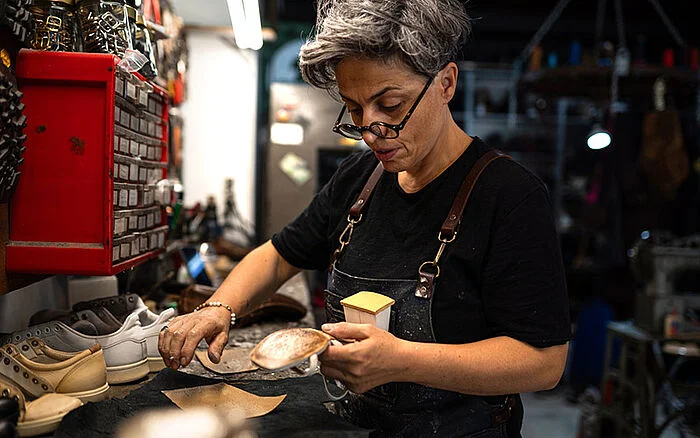 Shoemaker working at a cluttered workshop bench, polishing a leather shoe surrounded by tools and materials.