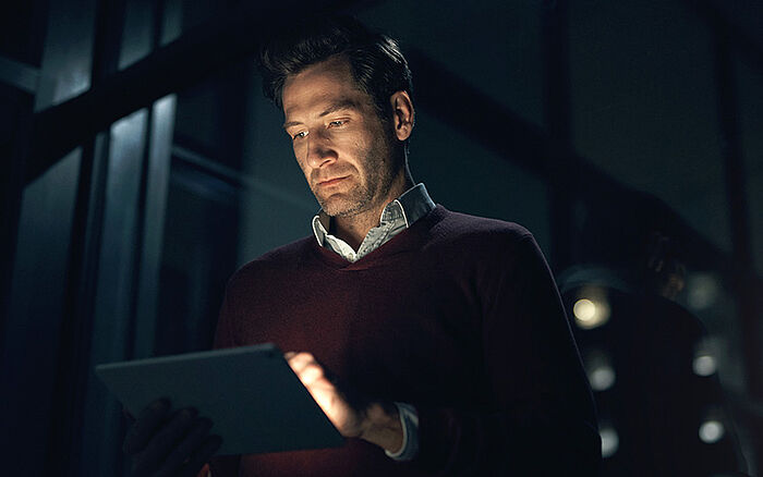 A man's face is lit up by the screen of his tablet as he reads the news with concern.