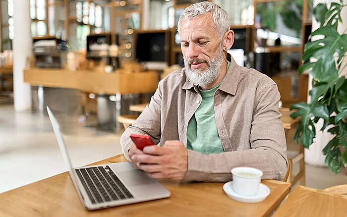 Middle-aged gray-haired businessman sitting at a café table, holding a smartphone and looking at the screen while working or texting.