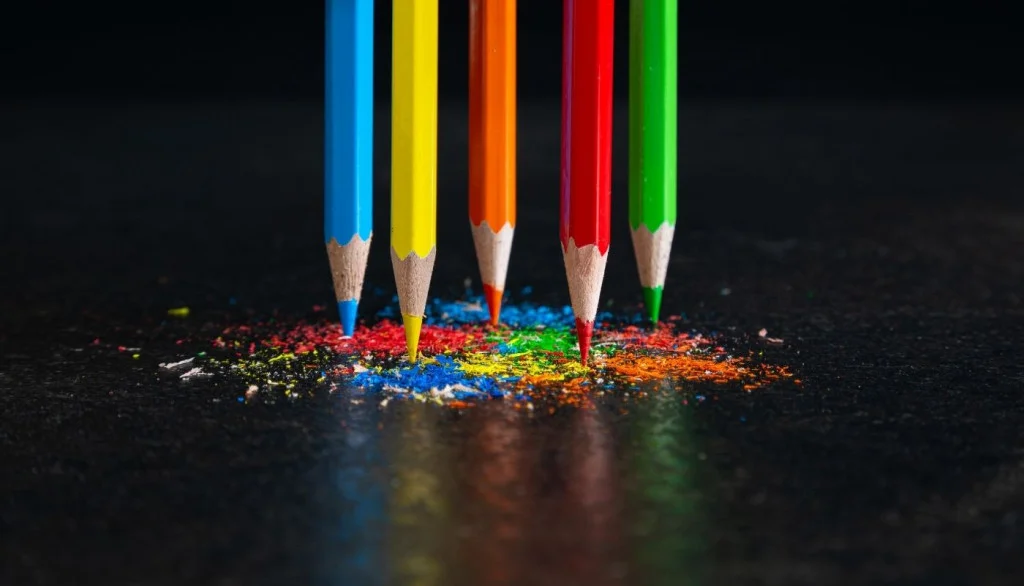 Five color pencils balance on their tips atop colored shavings on a black surface against a black background.