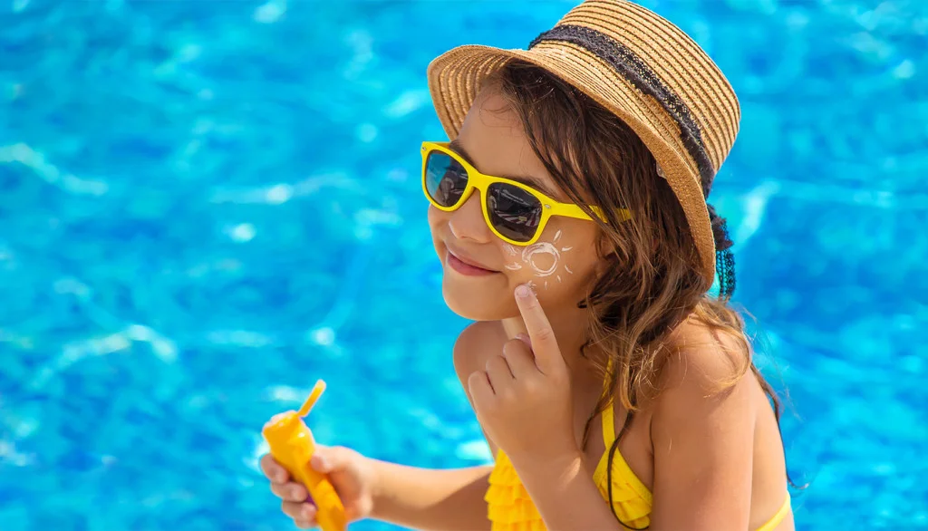 Young child applying sunscreen to her face with a focused expression, outdoors in natural light.
